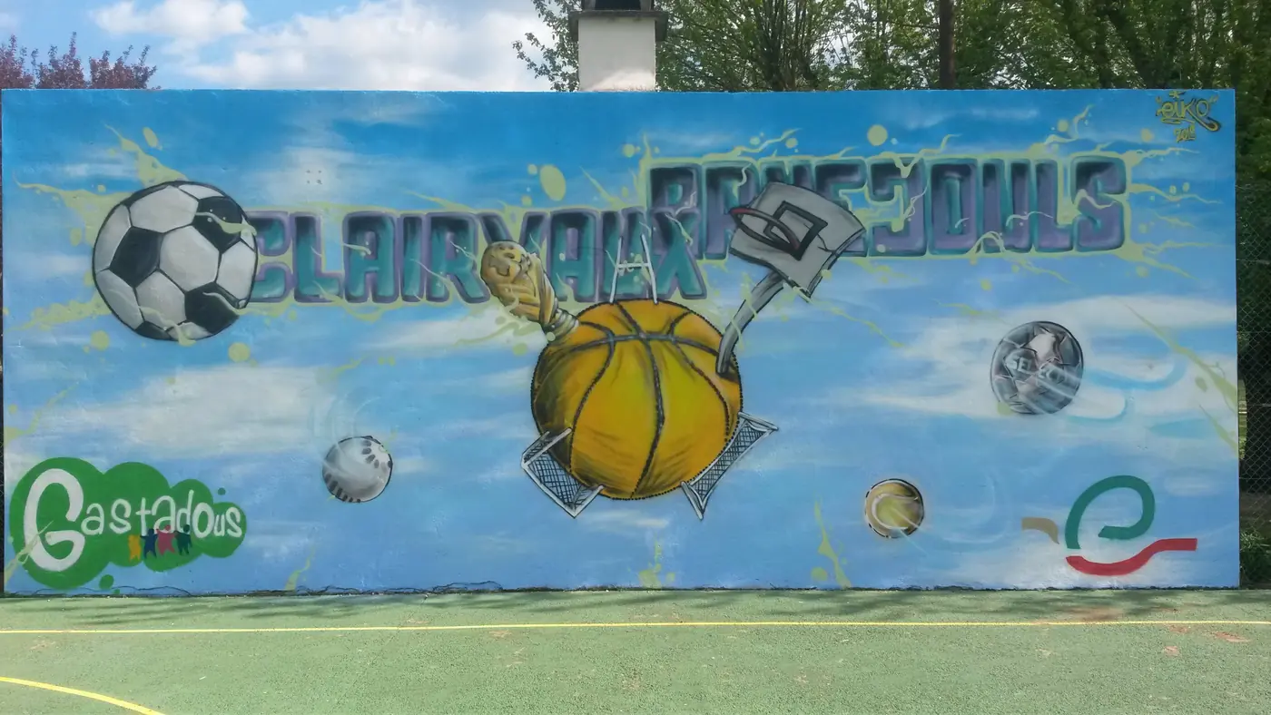 Fresque sur le mur du terrain de sport de Clairvaux-d'Aveyron : ballons de foot et de basket devant un ciel bleu